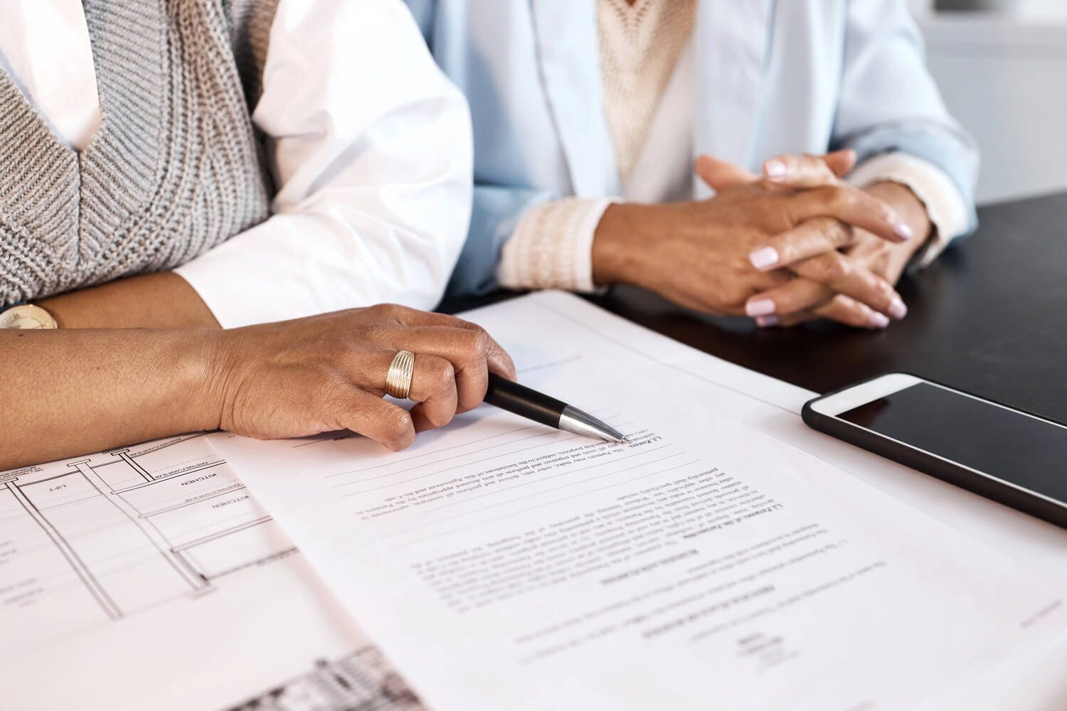 Two people are sitting down in front of a long legal document. The person on the left is holding a pen and using it to point at something in the document. The person to the right has their hands folded on the desk.