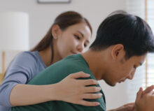 A young Asian female puts her arm around a young Asian man, who has his hands clasped and appears to be in grief