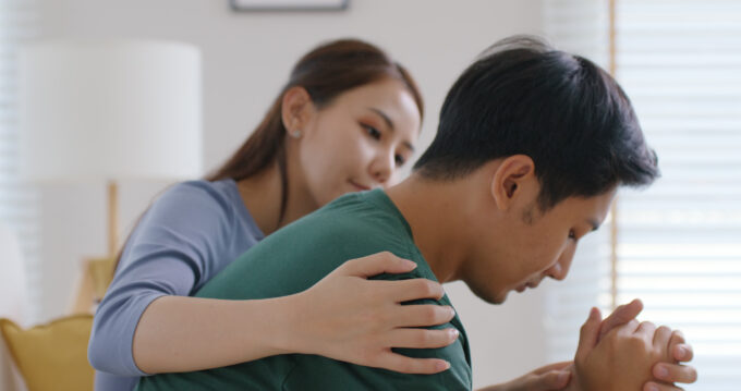 A young Asian female puts her arm around a young Asian man, who has his hands clasped and appears to be in grief