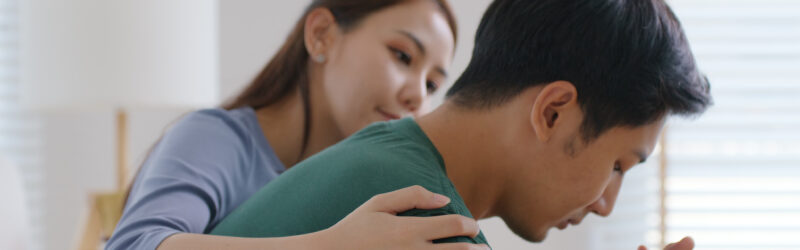 A young Asian female puts her arm around a young Asian man, who has his hands clasped and appears to be in grief